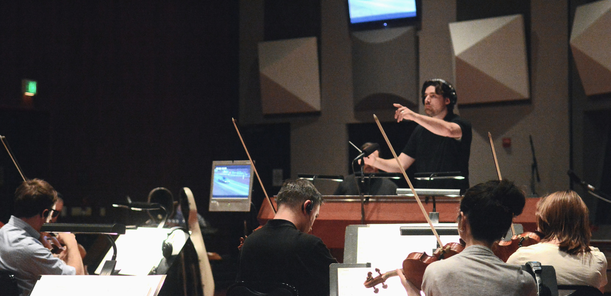 Composer, Jeff Toyne, conducting orchestra at Eastwood Scoring Stage, Warner Brothers Studios, Burbank, CA.  String section has their backs turned to camera. Pyramid shapes mounted on the studio wall behind Jeff. He wears dark short-sleeved shirt.