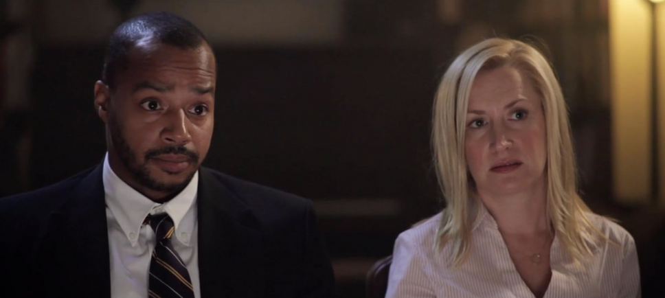 Scene from Hot Bot movie, starring Donald Faison and Angela Kinsey.  He is wearing a suit and striped tie with his eyebrows raised while she looks in the same direction in a light collared blouse.