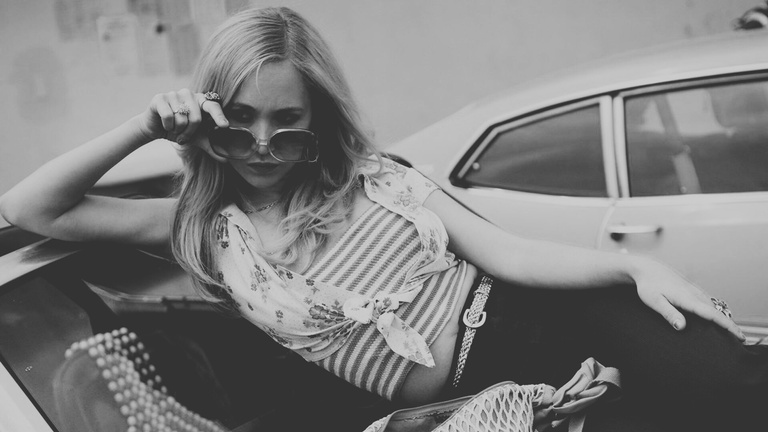 Black and white photo of a scene from feature film, Dirty Girl.  Juno Temple plays teenager, Danielle, lying on a car hood, seductively peering over her sunglasses. Click to view film dossier.