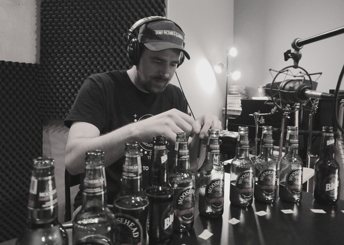 Composer Jeff Toyne in recording studio sampling beer bottle sounds by varying levels of beer. For A Year in Change movie.  He wears Sony Pictures Classic baseball cap and over-ear headphones. Panels of egg crate foam hang on the wall behind.