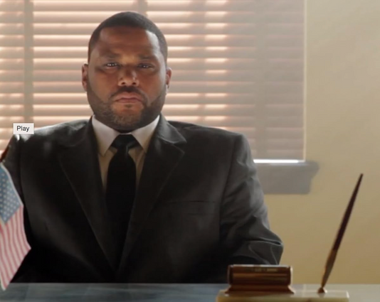 Agent Frazier played by Anthony Anderson sits upright and expressionless against a window with the blinds slightly open in a scene from Hot Bot.  He wears a dark suit, tie, and white collared shirt.