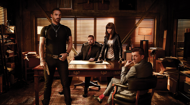 Cast photo for Rogue TV series in a dark detective's office with the blinds barely open. They sit and stand around a large wooden desk with piles of evidence around them. 