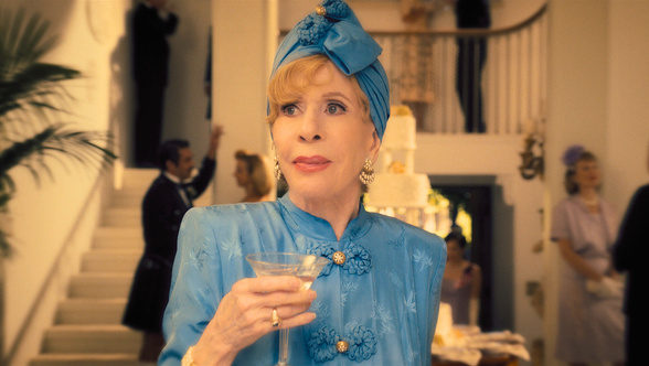 Carol Burnett plays Norma Dellacorte in Palm Royale. She is in the foreground of a glamorous party in her mansion.  Wearing a cornflower blue silk outfit with gold Chinese buttons and matching turban. She has a martini in hand.
