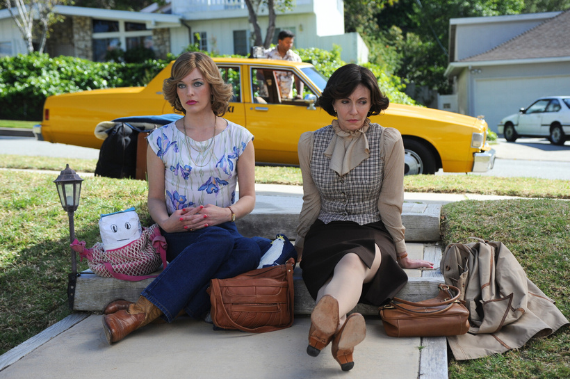Milla Jovovich and Mary Steenburgen's characters from Dirty Girl sit forlorn and defeated together on the lawn with an 80s yellow cab parked behind them.  They both have perfectly feathered bobs and typical 80s outfits on.