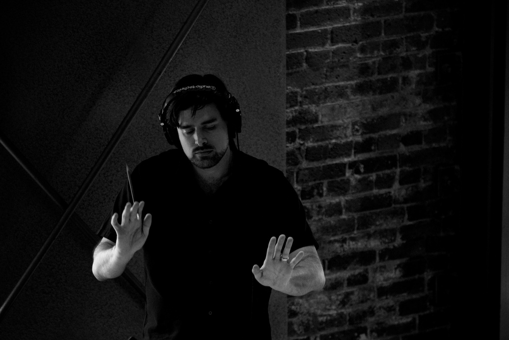 Medium shot of composer Jeff Toyne conducting, holding up baton with his other hand at same level, palm facing camera.  He is looking down, presumably at the score.  Wearing studio headphones and dark short sleeve shirt.  Brick wall behind him.