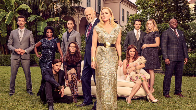 Filthy Rich cast photographed on the lawn in front of a mansion and tropical trees. Kim Cattrall at the center surrounded by 10 actors dressed in formal attire of their characters, including Gerald McRaney and Steve Harris.