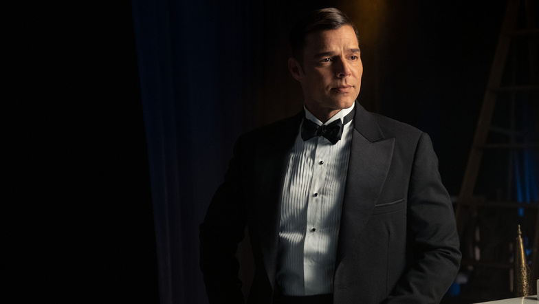 Photo of Ricky Martin playing Robert in Palm Royale, looking dapper and dramatic wearing a tuxedo against a dark background.  He has a somber expression looking off camera.