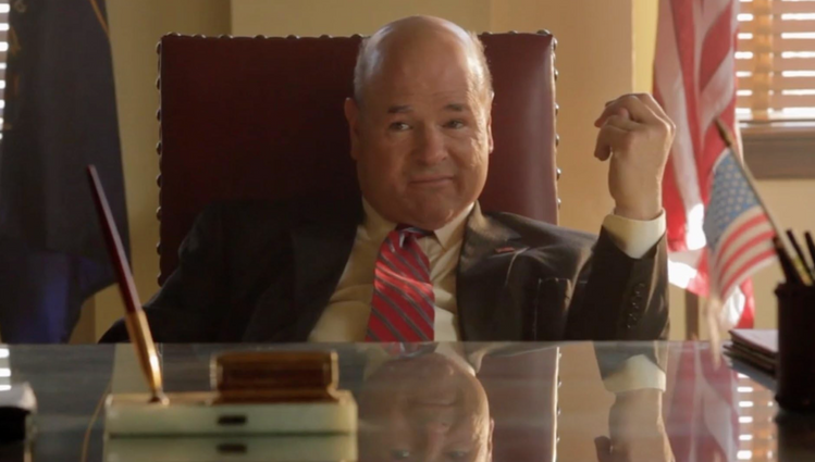 Senator Biter played by Larry Miller leaning back at his large leather chair at his glass-top desk.  There is an American flag raised on a stand behind him. He's wearing a red striped tie and suit. Typical politician's outfit.